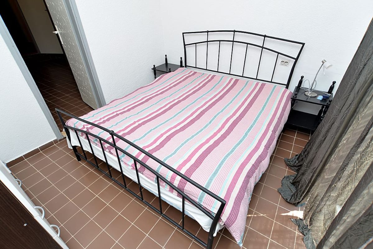 A single bed with a metal frame, a striped bedspread, a bedside table with a lamp, and a wall socket.