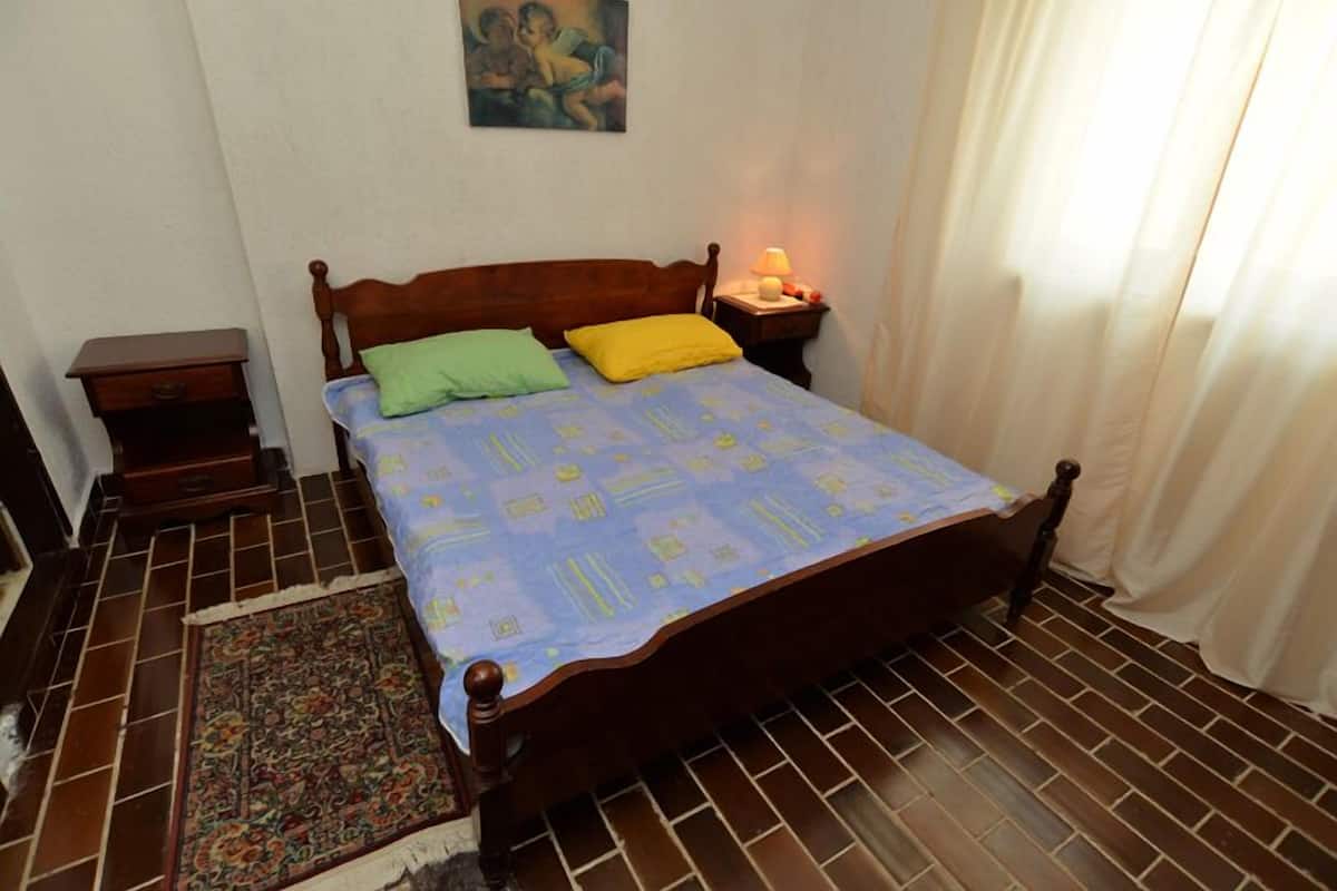 A bedroom with a wooden bed, a nightstand with a lamp, a patterned rug, and a painting on the wall.