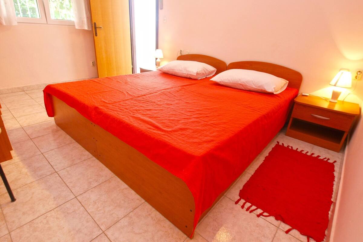 Quarto individual com cama de madeira, colcha vermelha, mesa de cabeceira com abajur e tapete vermelho.