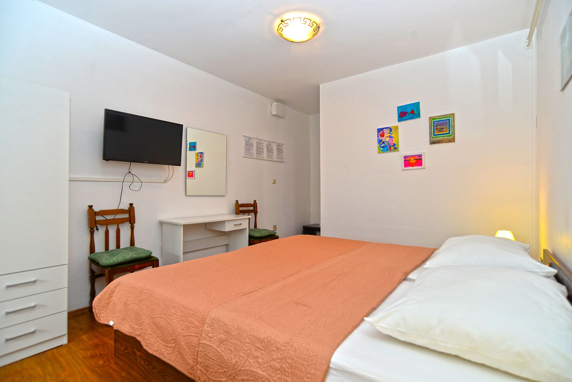 A bedroom with a bed, a desk, a chair, a television, and a wall with pictures.