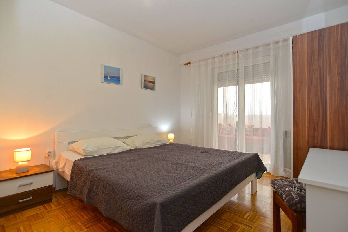 Quarto com cama, abajures, mesa de cabeceira de madeira, cadeira e janela com cortinas leves.