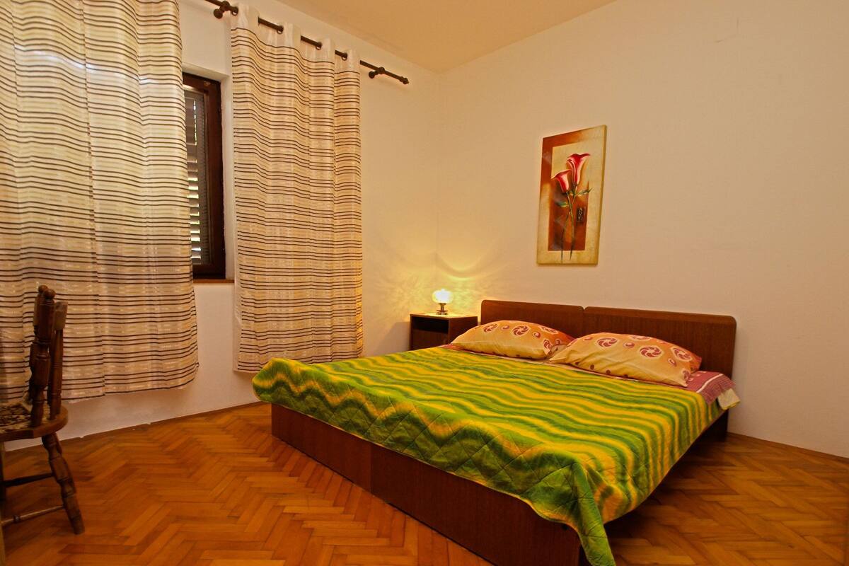 A bedroom with a wooden herringbone floor, a bed with a green and yellow patterned bedspread, a wooden headboard, and a nightstand with a lamp.