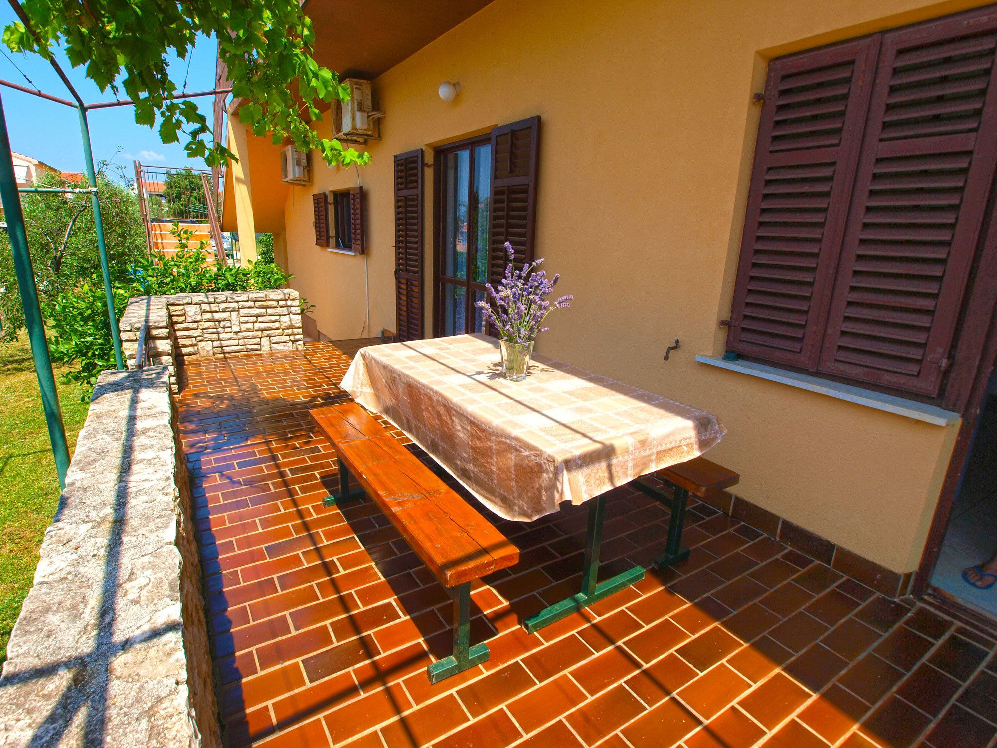 Une terrasse extérieure avec une table et des bancs, entourée d’un mur de briques et d’un jardin.