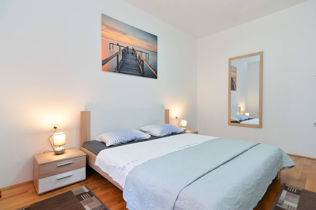 A bedroom with a bed, bedside tables, a nightstand, a wall-mounted mirror, and a painting of a pier.