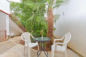 A small outdoor patio with two white plastic chairs and a round table, surrounded by lush greenery and a palm tree. - Apartment Kate 407 (Fazana)