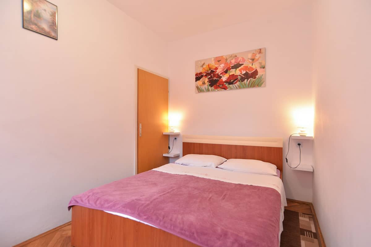 A bedroom with a wooden bed, white pillows, a pink blanket, and a floral painting on the wall.
