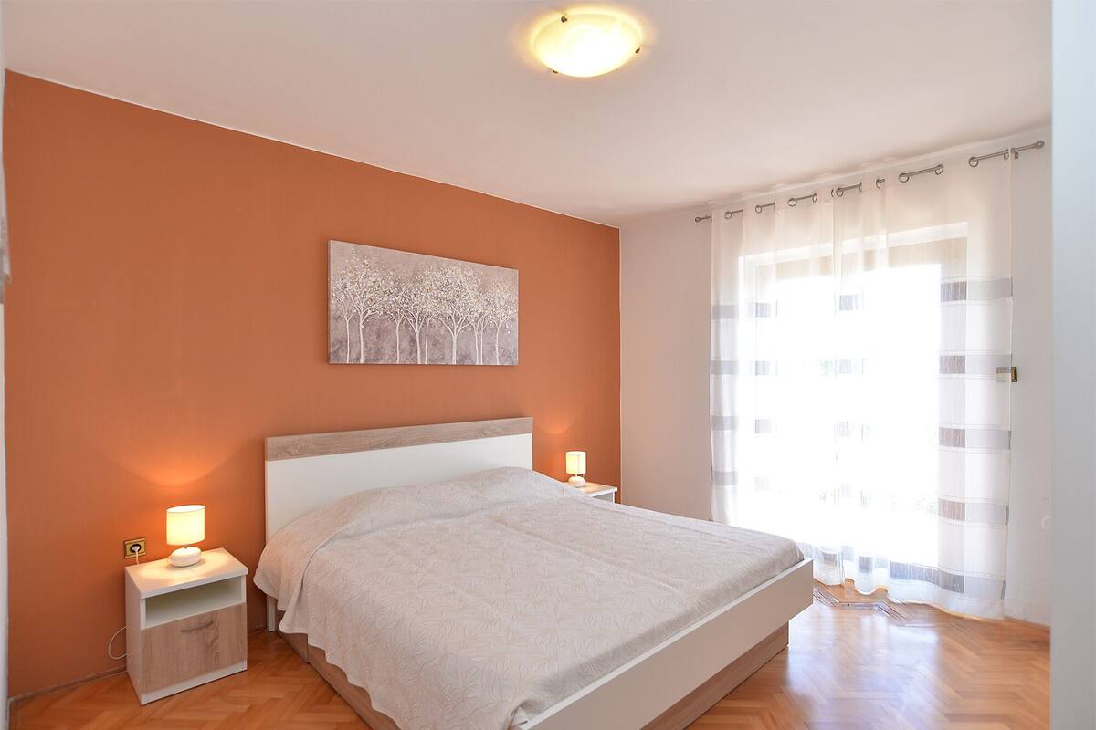 Un dormitorio con cama, mesitas de noche, una lámpara de pared, un cuadro y una ventana con cortinas.