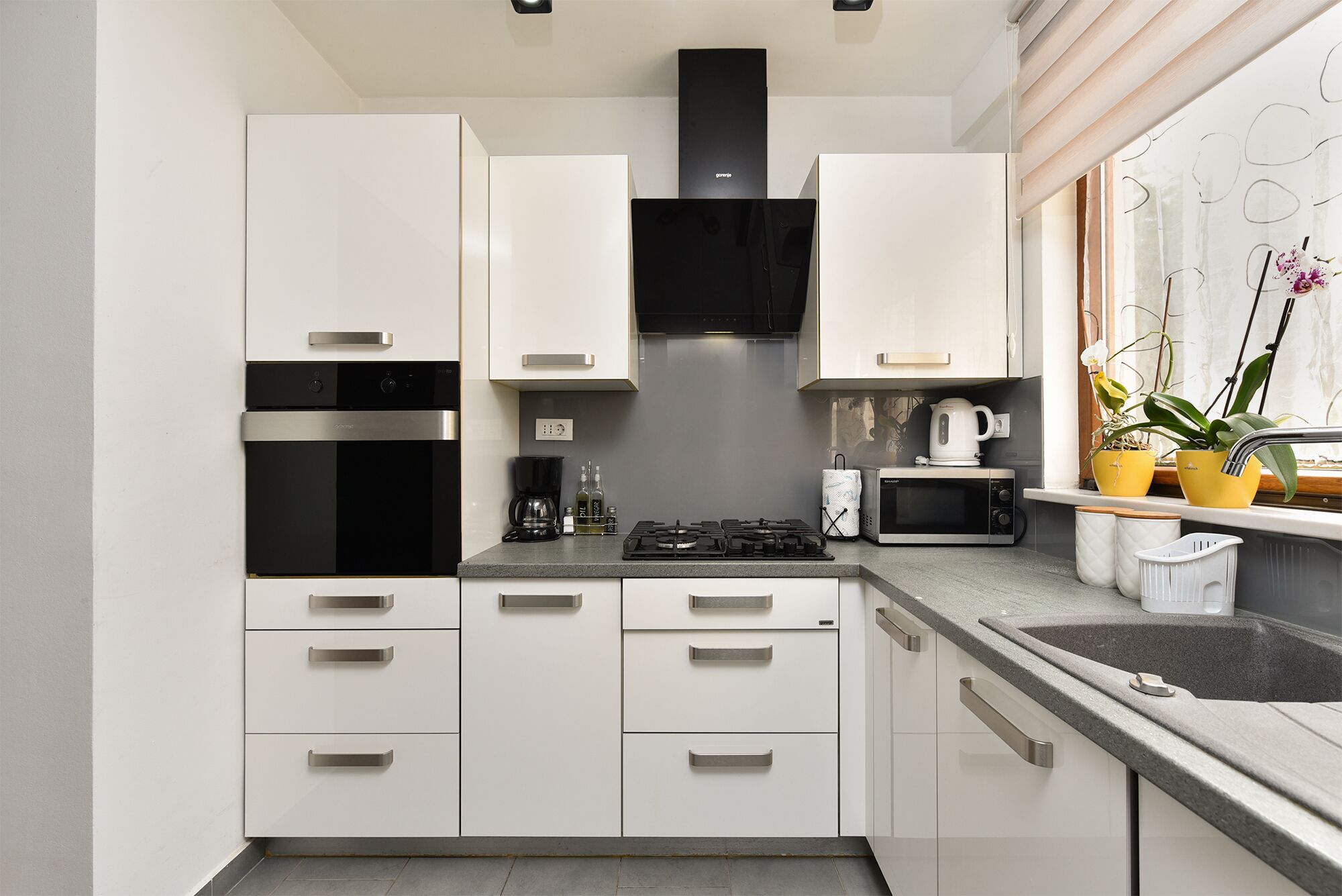 A modern kitchen with white cabinets, a black range hood, a microwave, a gas stove, and a window with plants.