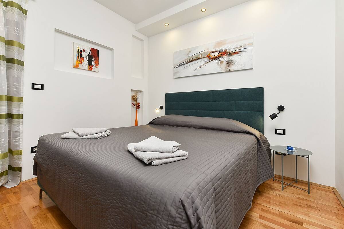 Un dormitorio moderno con una cama grande, dos obras de arte colgadas en la pared, una mesita de noche con una lámpara y suelo de madera.
