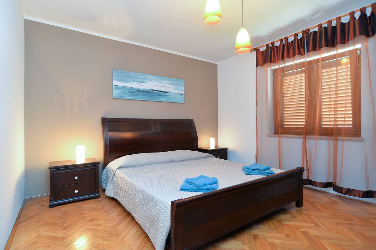 Un dormitorio con cama de madera, mesita de noche, un cuadro en la pared y ventana con persianas.