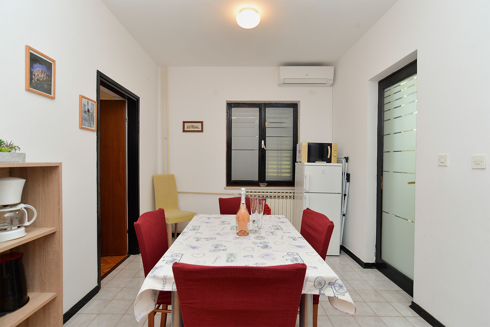 A compact dining area with a table set for four, surrounded by red chairs. A yellow chair is positioned against the wall, and a small cabinet with a plant is visible on the left.