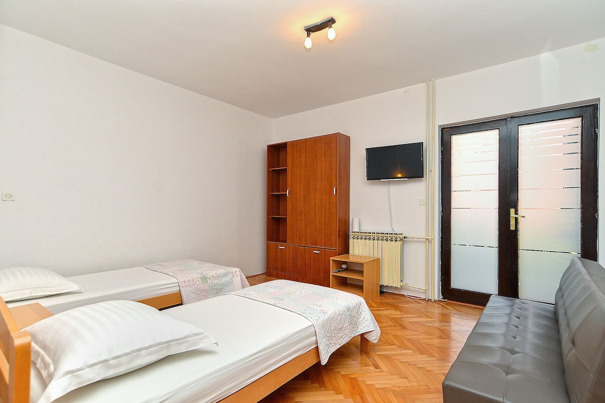 Quarto com duas camas de solteiro, guarda-roupa de madeira, televisão fixada na parede e porta de vidro deslizante para uma varanda.