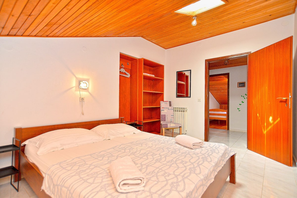 Um quarto com teto de madeira, uma cama com colcha estampada, uma mesinha de cabeceira, um guarda-roupa e um espelho na parede.