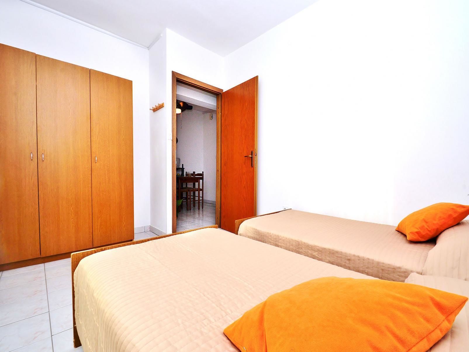 A bedroom with two beds, wooden wardrobes, and a dining area visible through an open door.