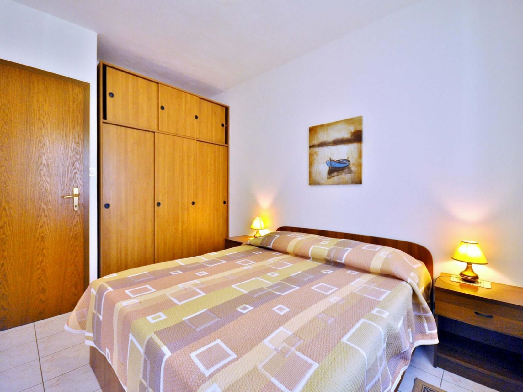 Quarto com cama, guarda-roupa de madeira, duas luminárias de cabeceira e um quadro na parede.