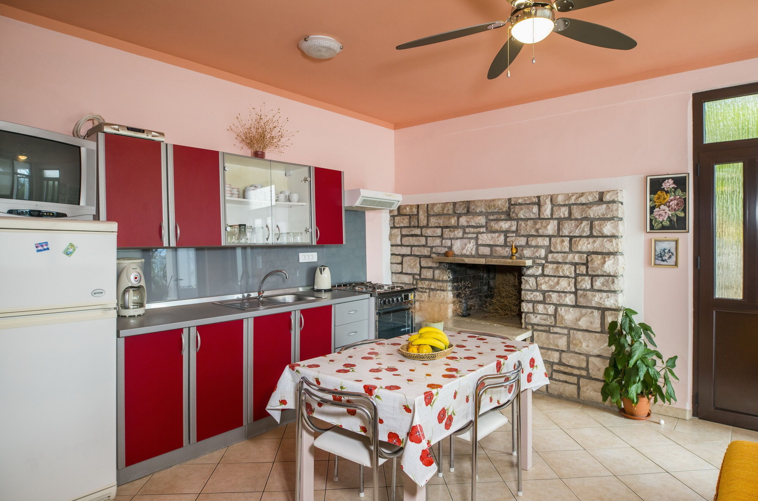 A kitchen with red cabinets, a stone fireplace, and a dining table with a fruit bowl.