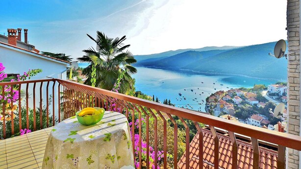 Ein Balkon mit Tisch, Obstschale und Blick auf eine Küstenstadt mit Booten und Bergen.