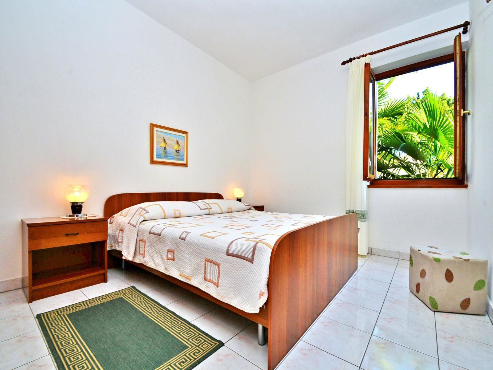 Une chambre à coucher comprenant un lit, des tables de chevet, une commode, une fenêtre donnant sur un paysage verdoyant et un tableau encadré accroché au mur.