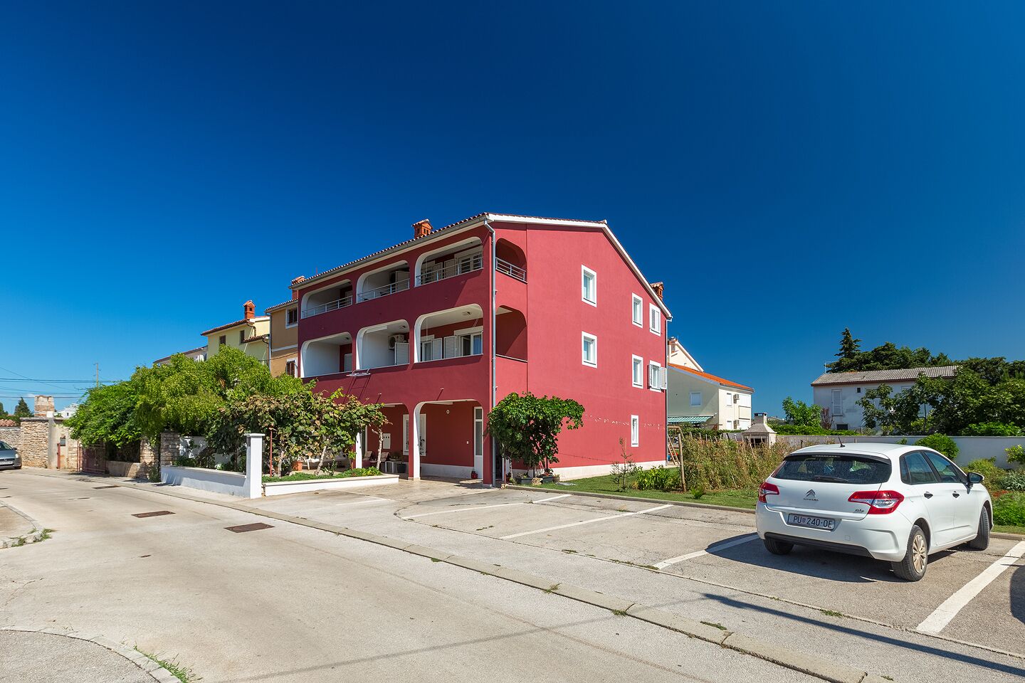 A red multi-story residential building with balconies, a parking area, and a white car parked in front.