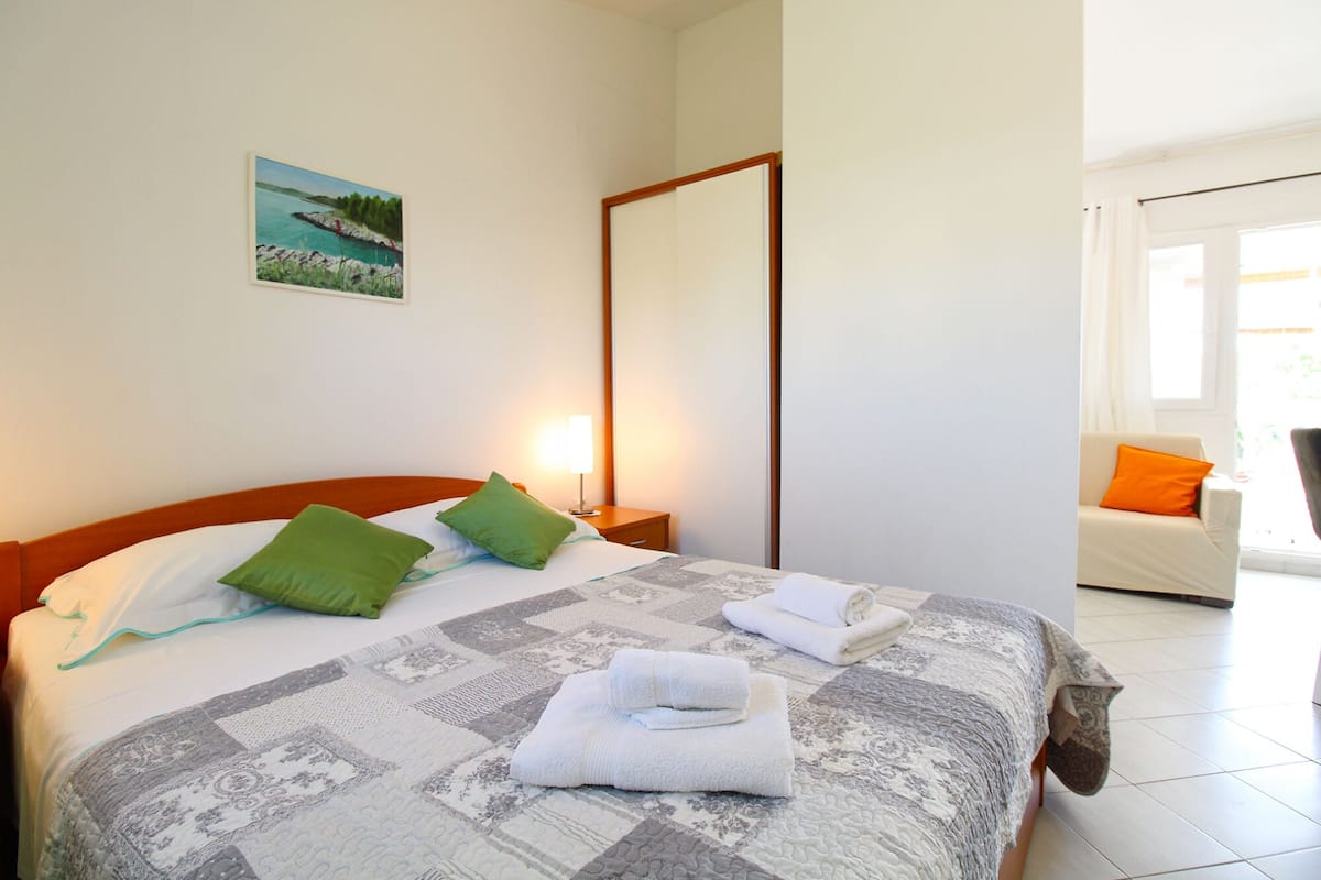 A hotel room with a bed, two pillows, a nightstand, a lamp, a painting, a mirror, a sofa, and a window.