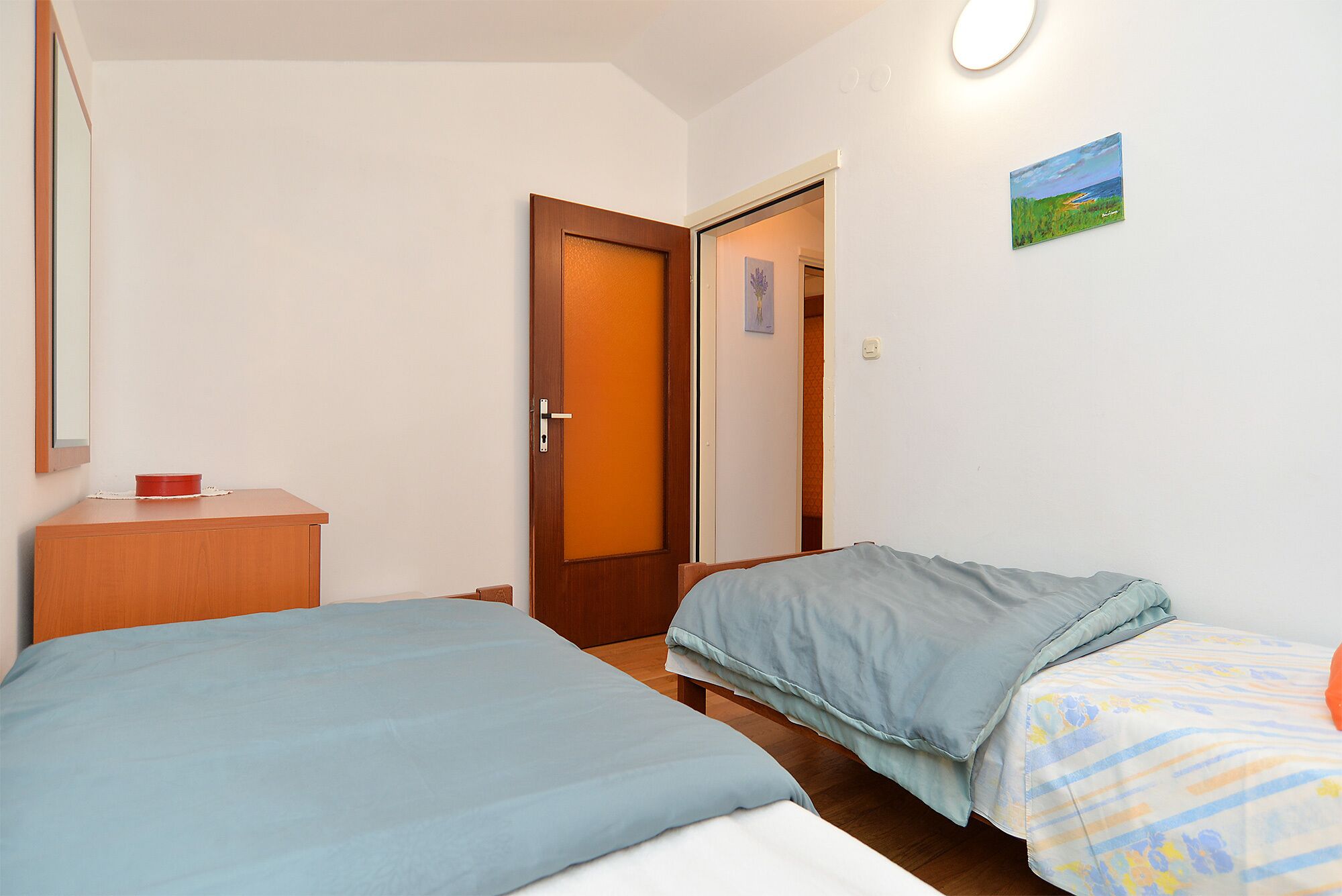 Une chambre avec deux lits, une commode en bois, une porte et un tableau accroché au mur.
