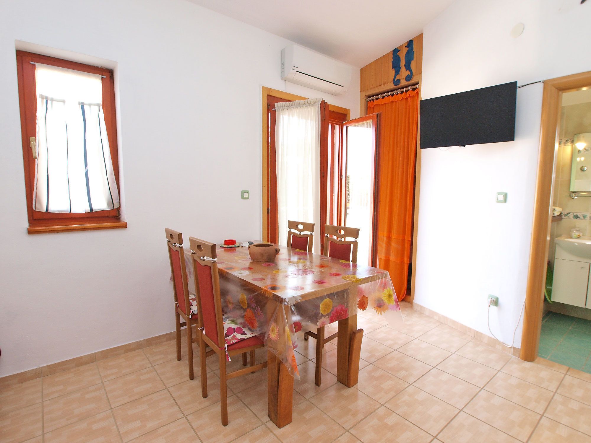 A dining area with a table covered in a floral tablecloth, surrounded by wooden chairs. There is a window with curtains, an air conditioning unit on the wall, and a television mounted above the table.