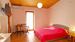 Un dormitorio con techo de madera, una cama con manta roja, una mesita con una silla y un televisor montado en la pared.