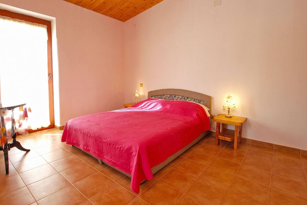 A bedroom with a wooden ceiling, a bed with a red blanket, two bedside tables with lamps, and a window with curtains.