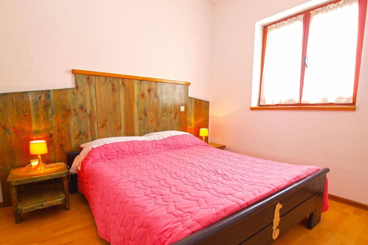 A bedroom with a wooden headboard, a pink quilted bedspread, two bedside lamps, and a window with curtains.