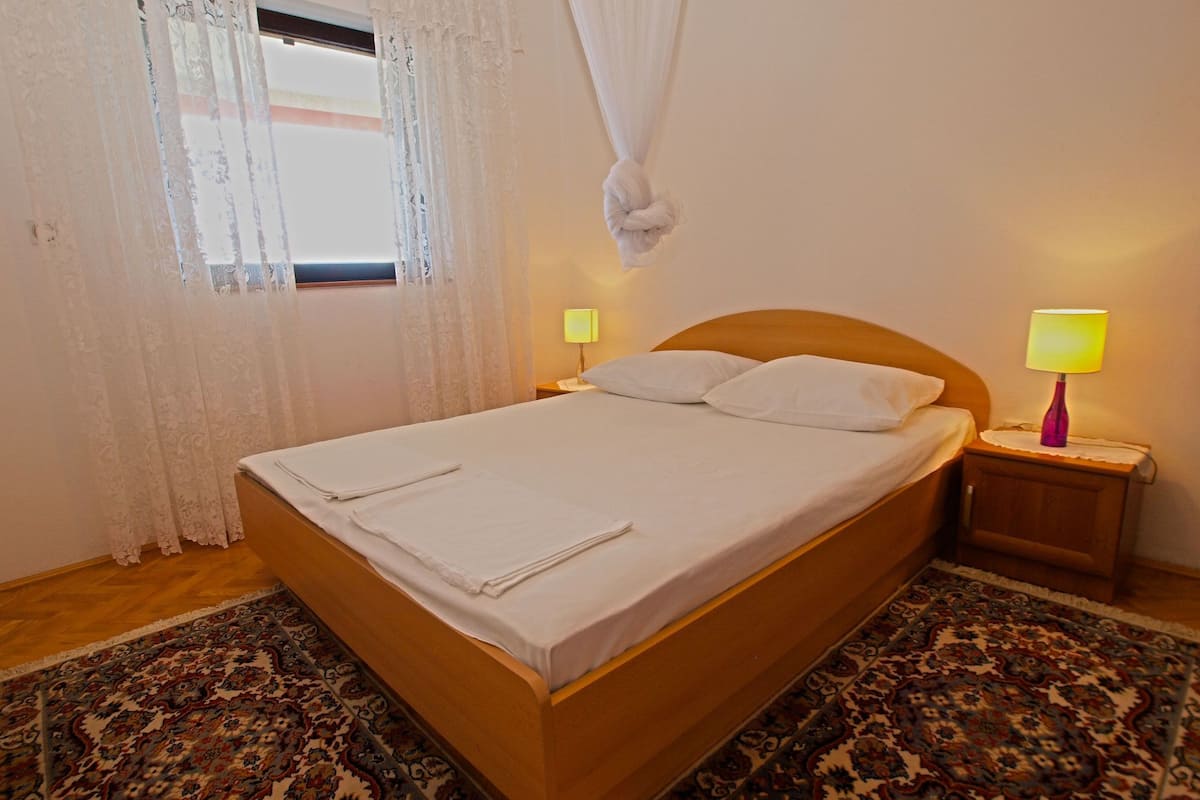 A neatly made bed with white linens, a wooden headboard, two bedside lamps, and a patterned rug.