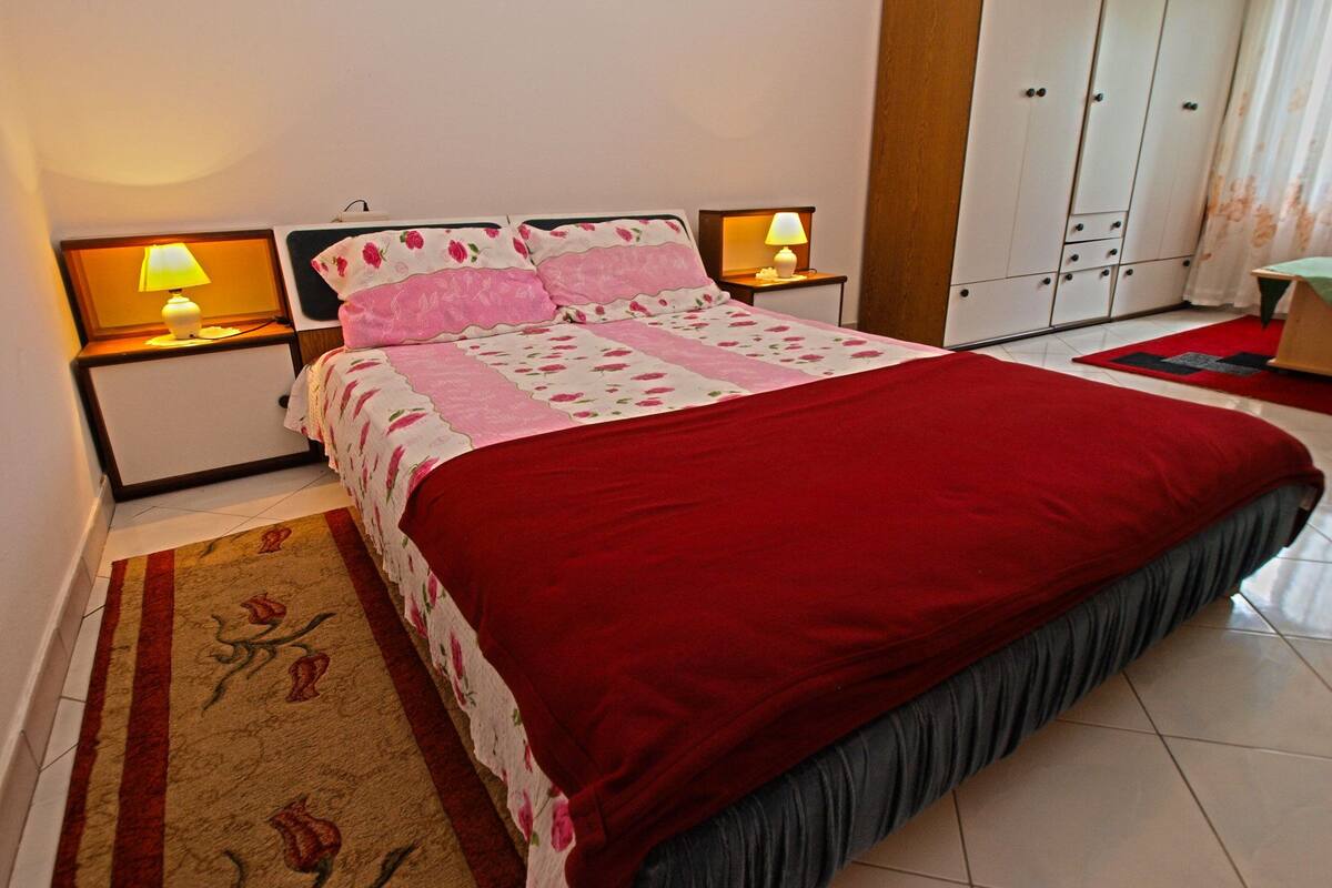 A neatly made bed with a red cover, two bedside lamps, a floral-patterned bedspread, and a red and beige rug with a rose design.