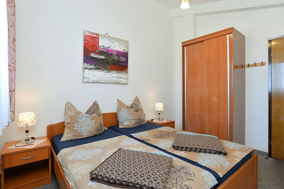A double bed with patterned bedding, two bedside tables with lamps, a wooden wardrobe, and a wall-mounted abstract painting.