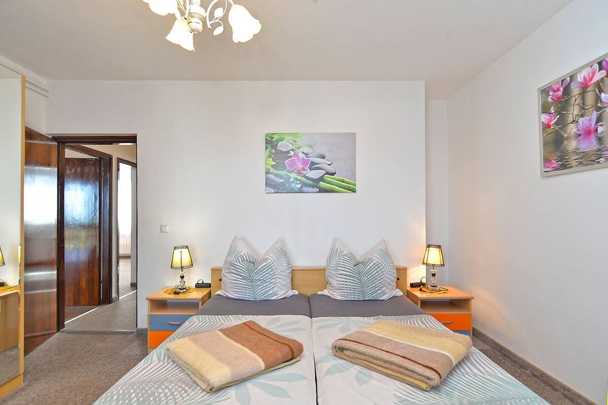 A hotel room with a bed, two bedside tables, a nightstand, a wooden door, and a painting of flowers on the wall.
