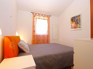 Ein Schlafzimmer mit Bett, Nachttisch, Lampe, einem Fenster mit Vorhängen und einem Bild an der Wand.