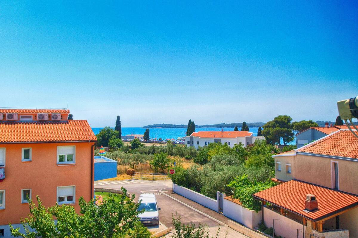 A coastal residential area with terracotta-roofed houses, a clear blue sky, and a view of the sea.