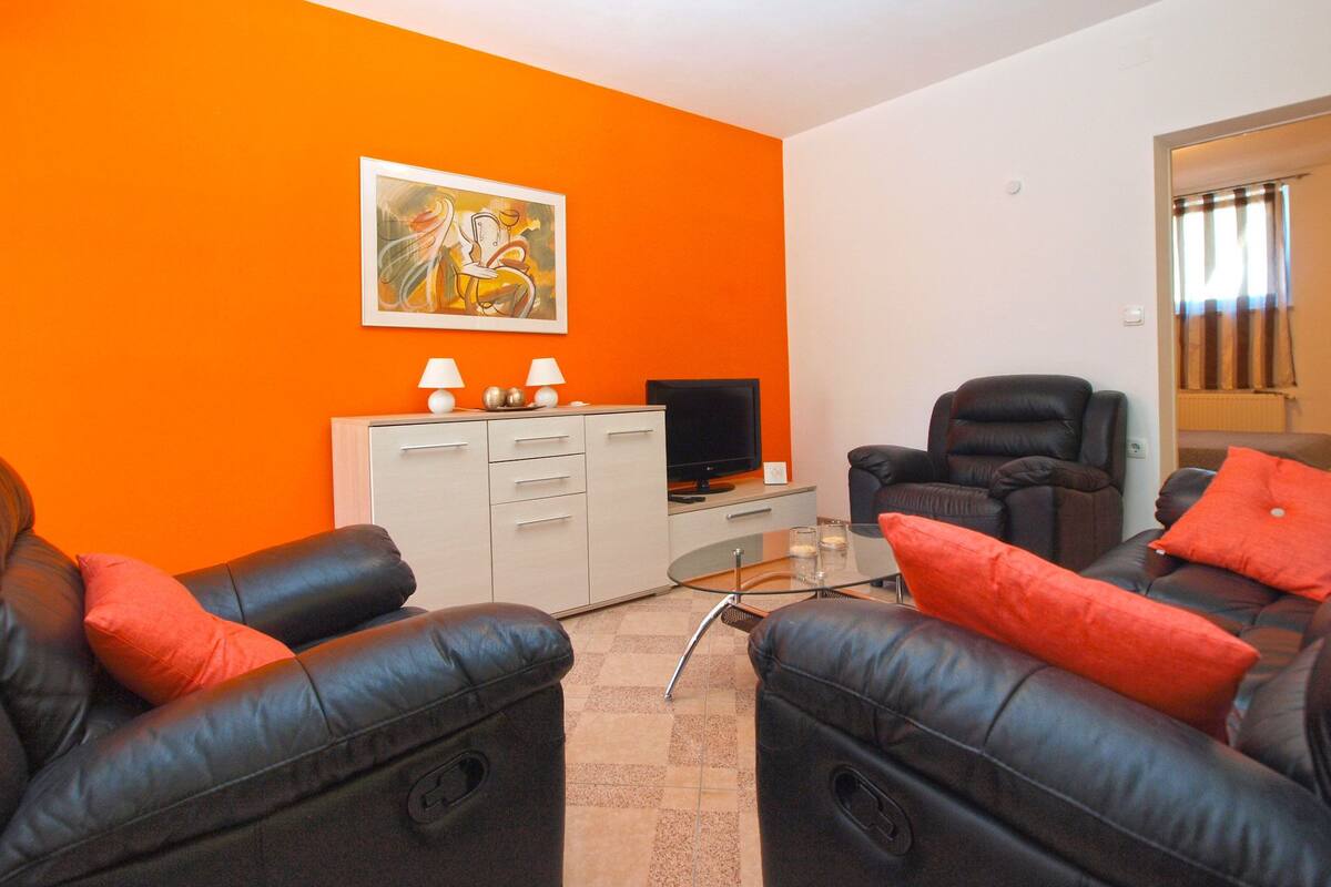 A living room with orange walls, a black leather sofa, a recliner, a TV, and a cabinet with two lamps.
