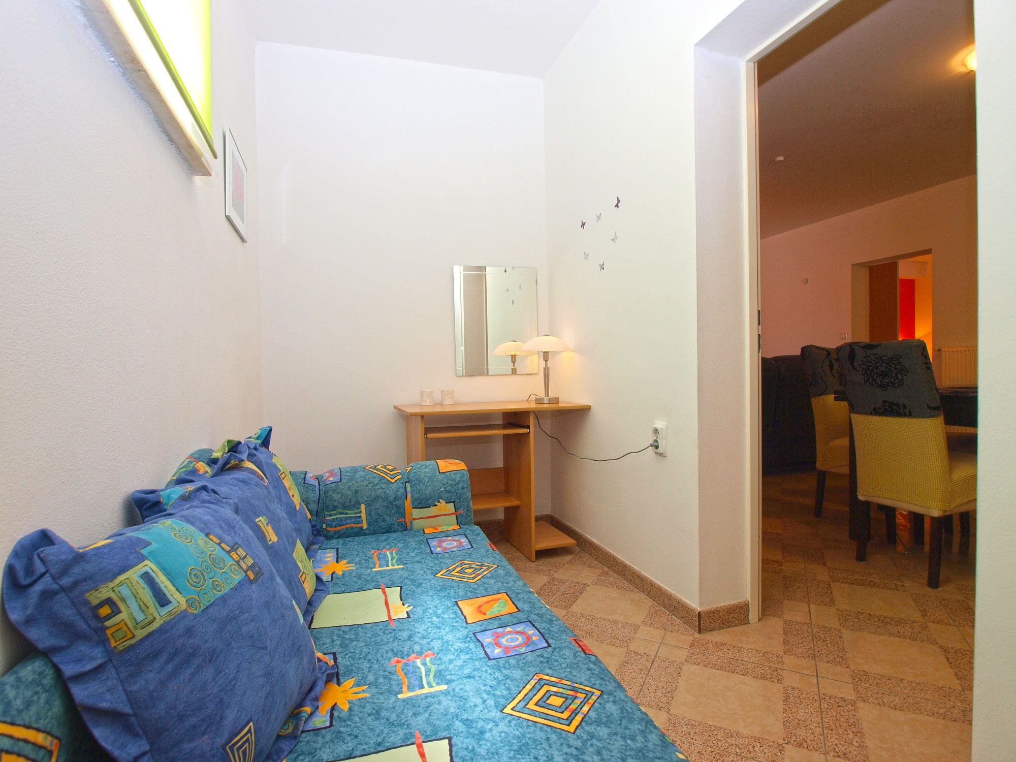 A small, cozy room with a bed, a desk, and a chair. There is a mirror above the desk and a small table with a lamp. The room has a tiled floor and a door leading to another room.
