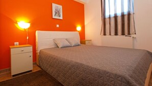A bedroom with an orange wall, a bed with a quilted cover, two bedside lamps, a nightstand, a radiator, and a window with curtains. - Apartments Ankica 1420 (Fazana)