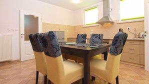 A dining area with a table and chairs, a kitchen in the background, and a window with blinds. - Apartments Ankica 1420 (Fazana)