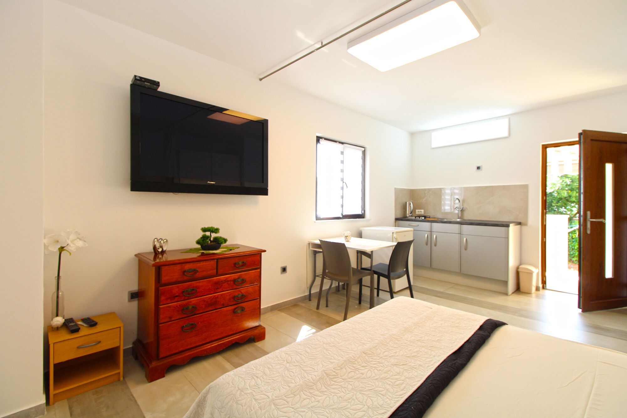 A bedroom with a bed, a dresser, a desk with chairs, a television, and a kitchenette.
