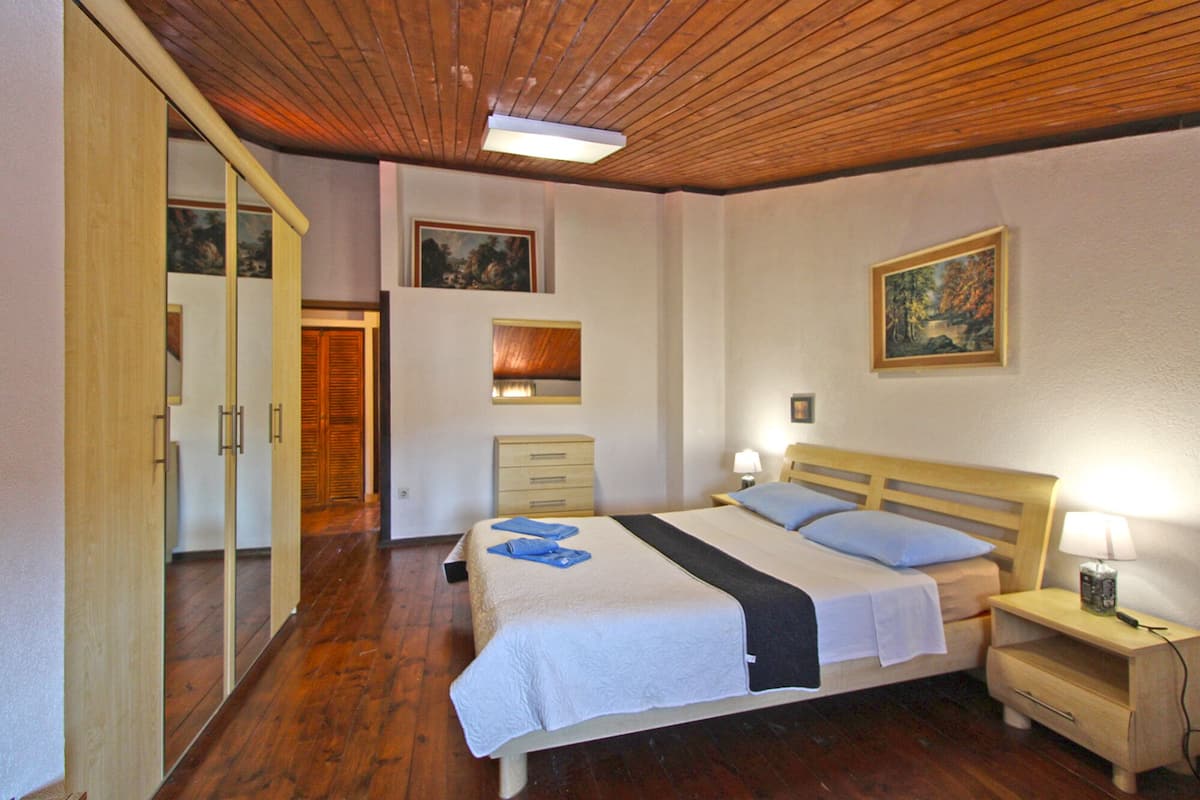 A bedroom with a wooden ceiling, a bed with a white and blue bedspread, a bedside table with a lamp, and a wardrobe.