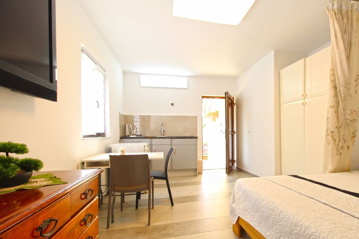 A modern bedroom with a bed, a desk, and a chair. There is a small kitchen area with a sink and a window.