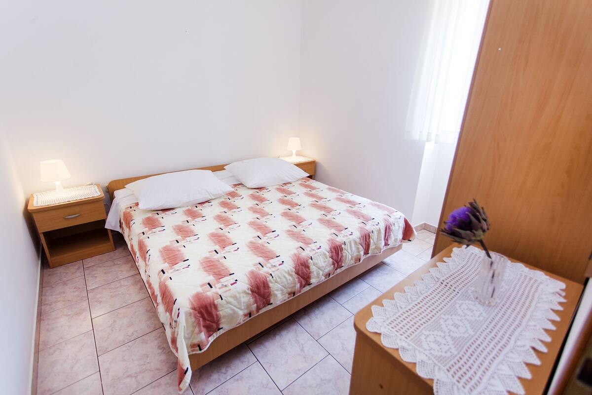 Quarto com cama, duas mesas de cabeceira com abajures, guarda-roupa de madeira e uma pequena mesa com um objeto decorativo.