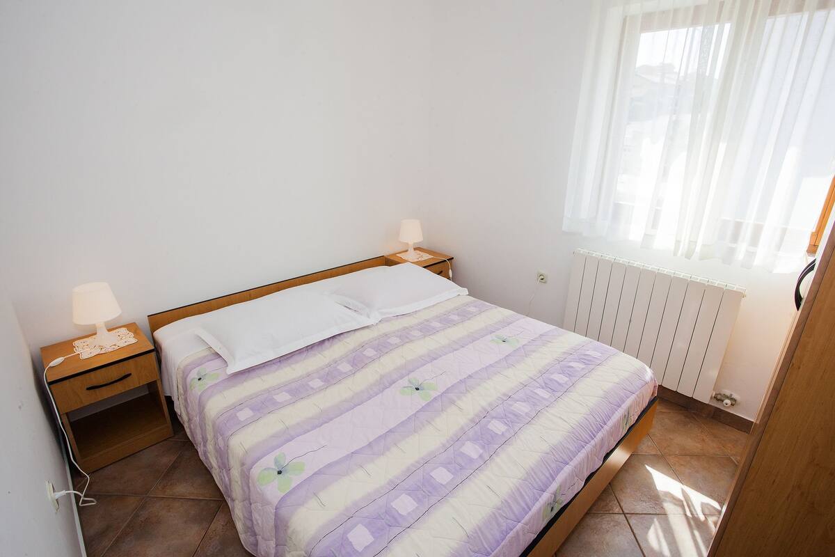 Quarto com cama, duas mesinhas de cabeceira, janela com cortinas leves e radiador.
