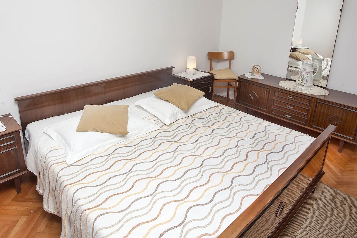 A neatly made bed with a patterned bedspread, a wooden nightstand with a lamp, a dresser with drawers, and a chair.