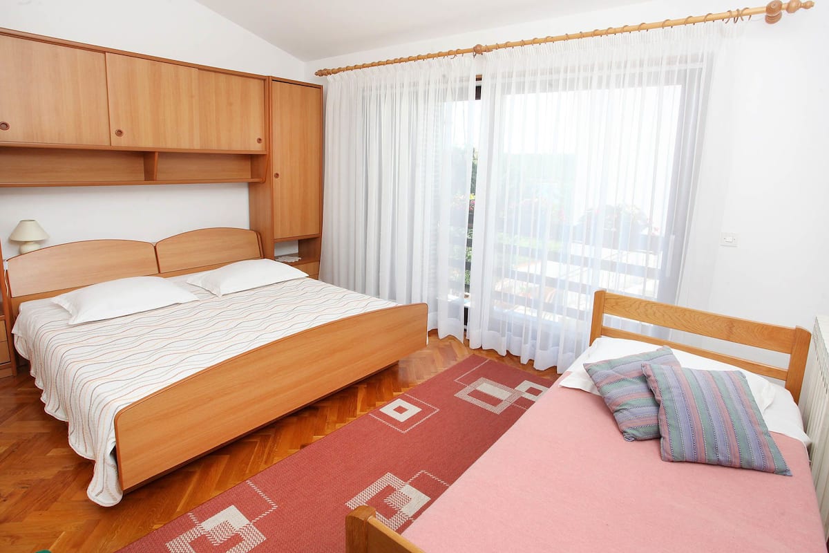 A bedroom with two beds, wooden headboards, a wooden wardrobe, a window with sheer curtains, and a wooden floor.