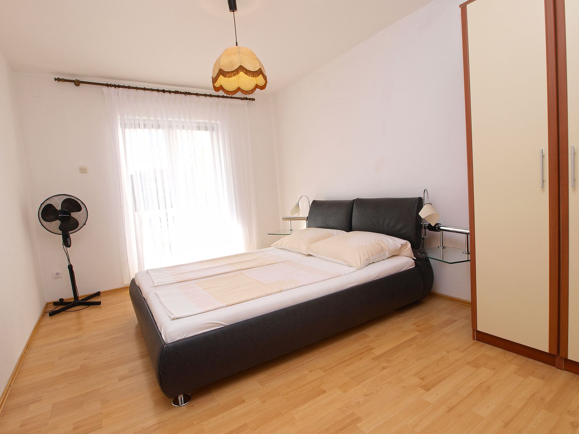 A bedroom with a bed, bedside table, lamp, and a standing fan.