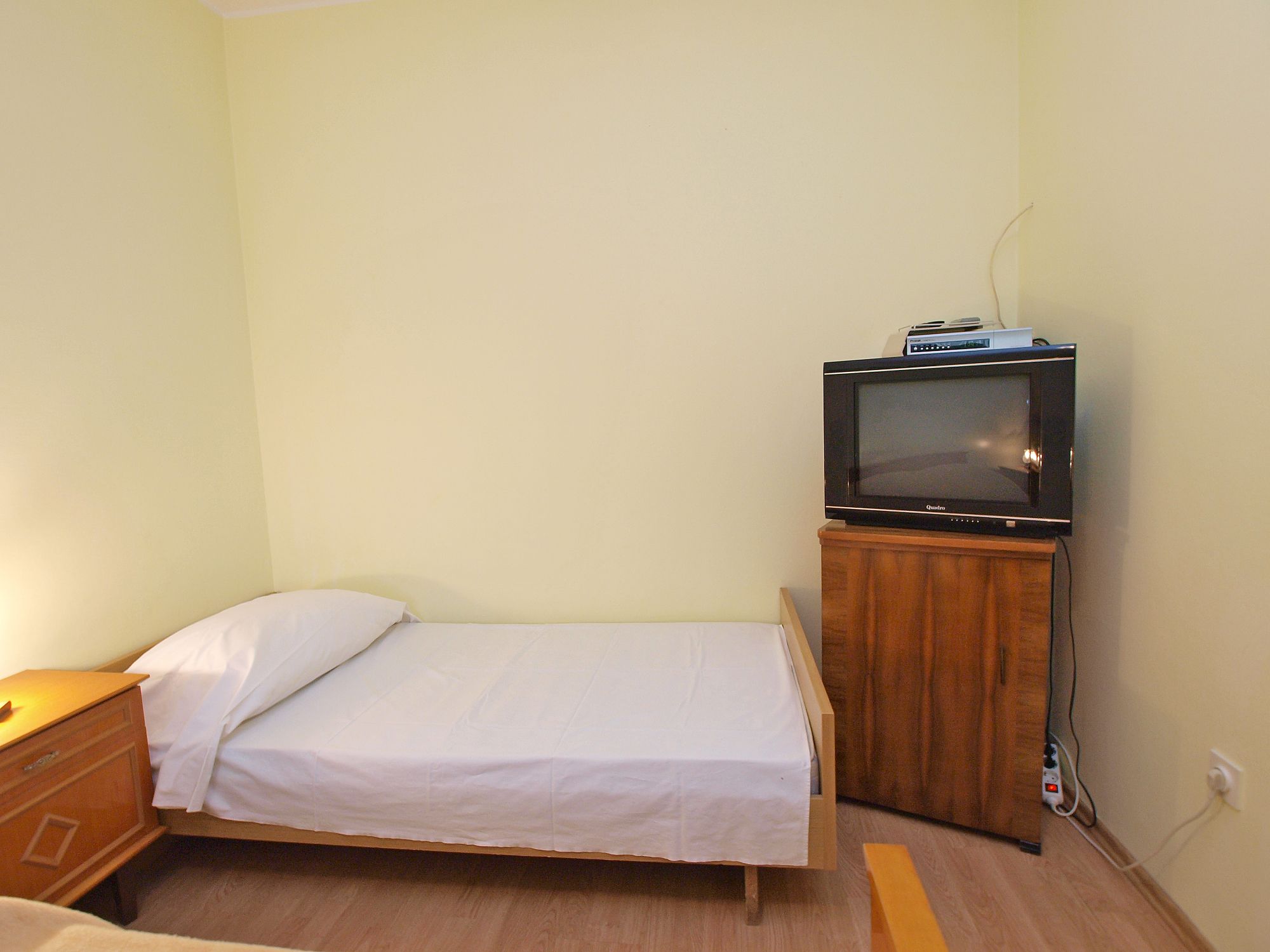 A small, single-bed room with a wooden nightstand, a television on a wooden cabinet, and a power outlet on the wall.