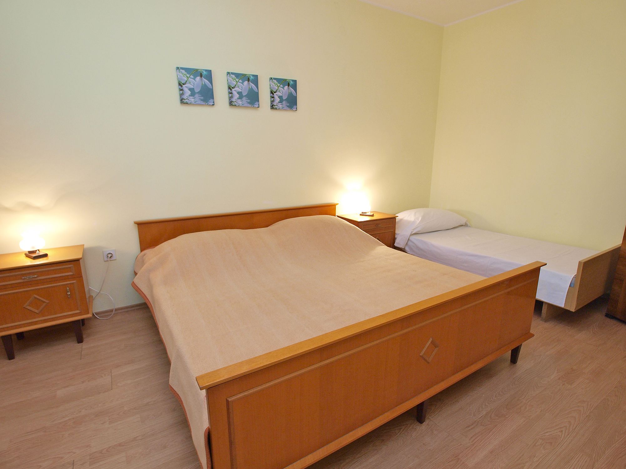 Een slaapkamer met een houten bed, een nachtkastje en een klein nachtkastje met een lamp. Er hangen drie ingekaderde foto’s aan de muur.