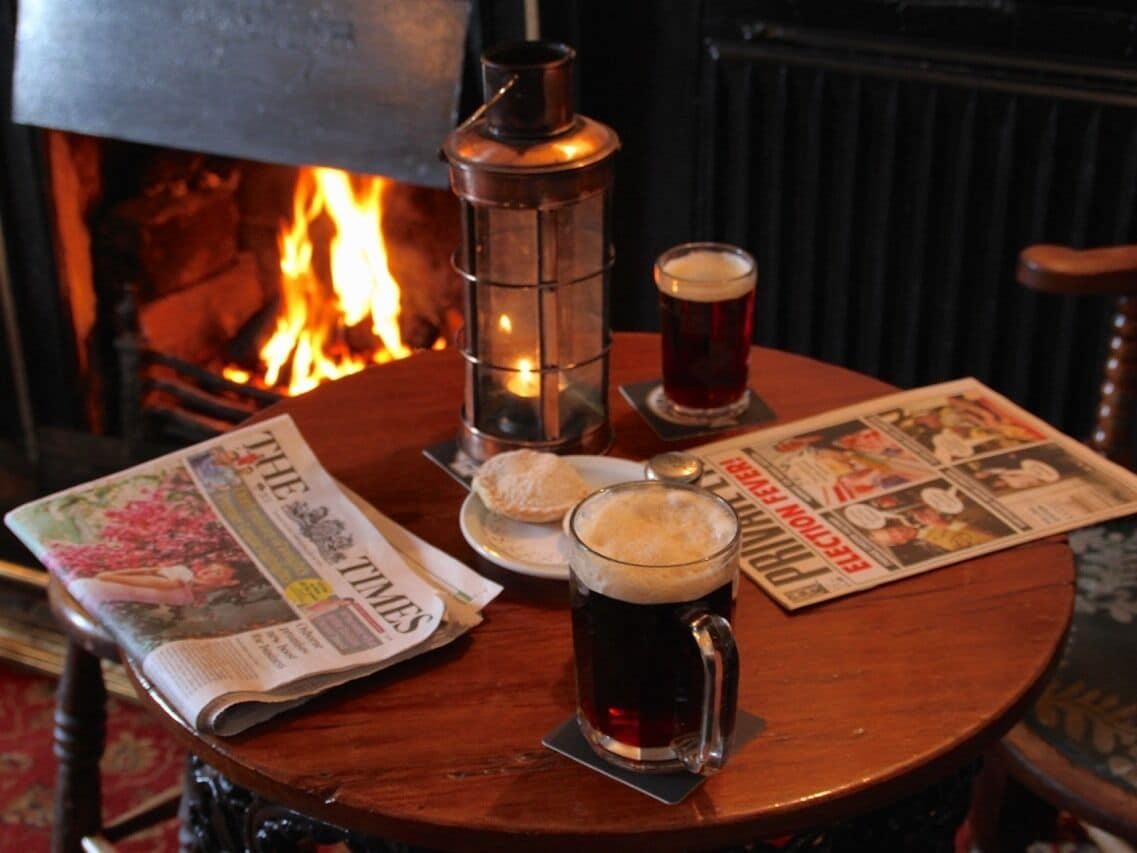 A table with a newspaper, a mug of beer, a glass of beer, and a candle holder.